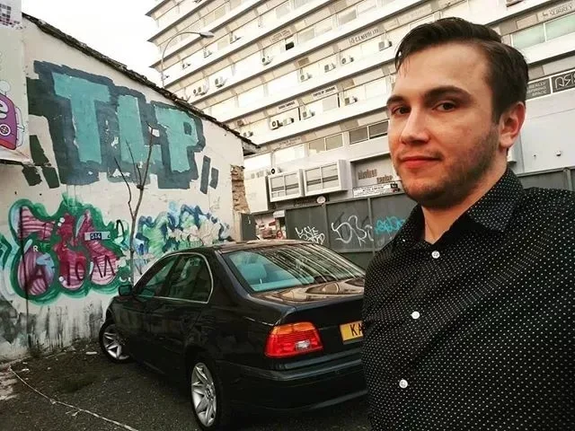 Андрей Чепелев рядом с чёрным автомобилем и граффити в Лимассоле