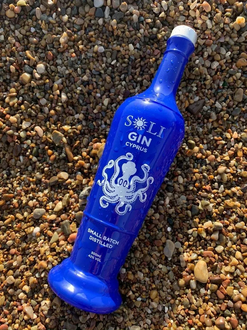 Бирюзовая бутылка Soli Gin лежит на гальке у берега с видимым логотипом