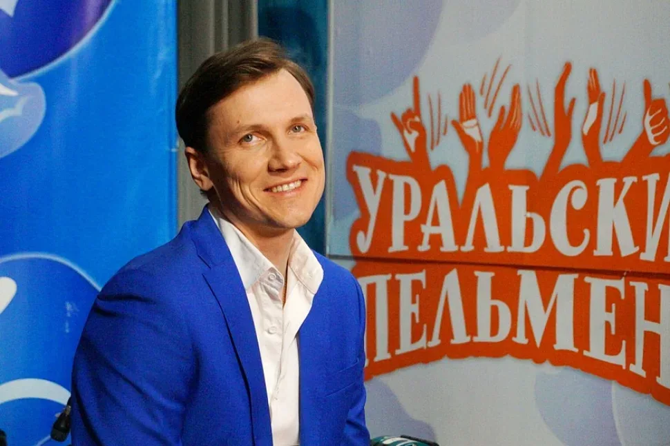Улыбающийся мужчина в синем пиджаке на фоне логотипа «Уральские пельмени