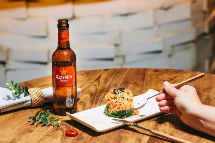 Бутылка Estrella Damm рядом с тарелкой тапас и рукой с вилкой