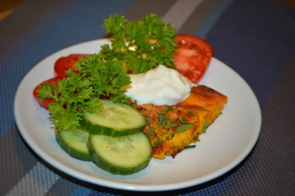 Порция морковной запеканки на тарелке с огурцами, помидорами и сметаной