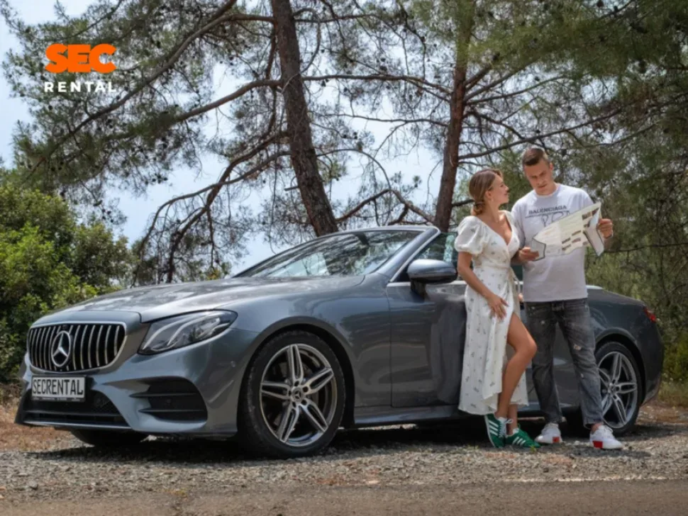 Пара у арендованного кабриолета Mercedes рассматривает карту среди сосен Пара у арендованного кабриолета Mercedes рассматривает карту среди сосен