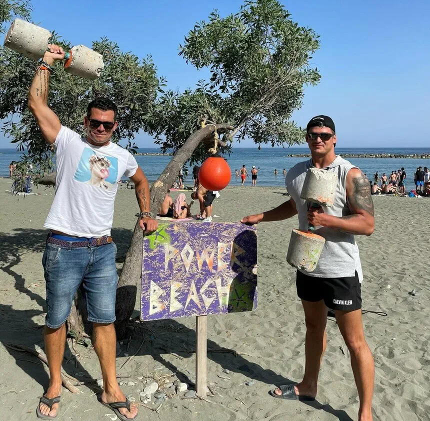 Два мужчины рядом с табличкой Power Beach на пляжном берегу