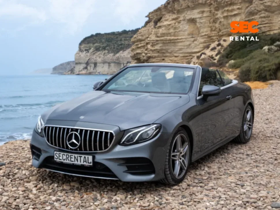 Серый кабриолет Mercedes с табличкой Secrental на галечном побережье у скал Серый кабриолет Mercedes с табличкой Secrental на галечном побережье у скал