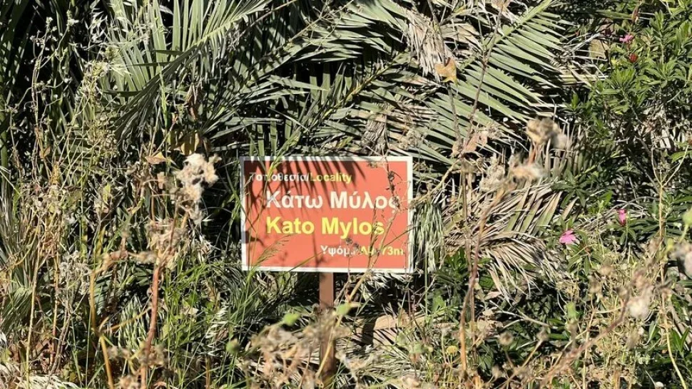 Туристическая табличка Κάτω Μύλος (Kato Mylos) на фоне растений