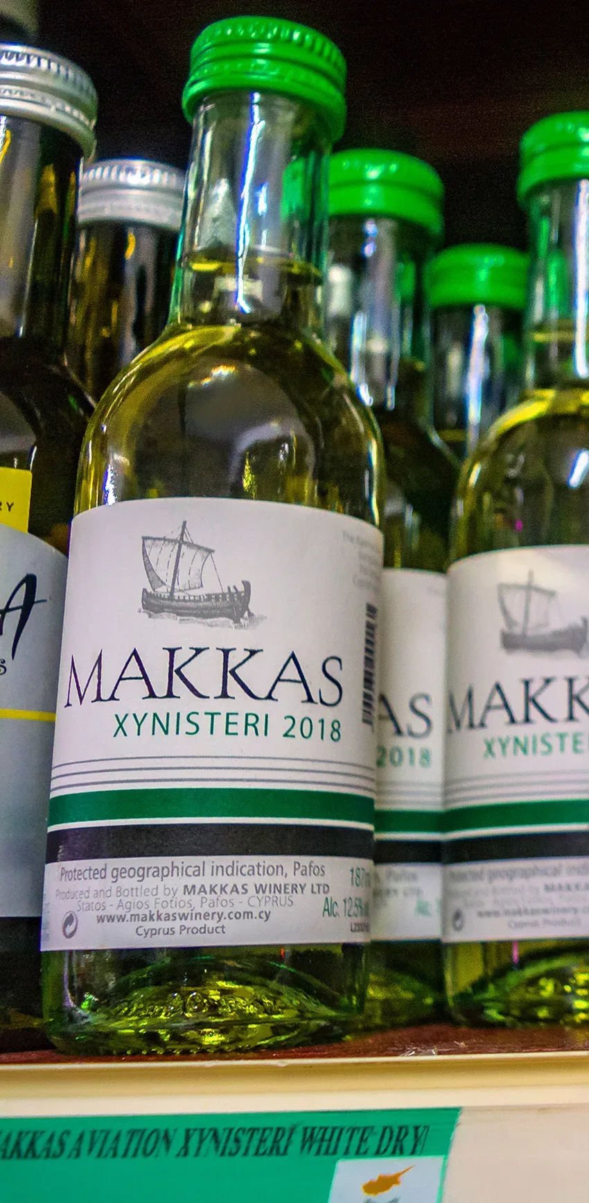 Бутылка Makkas Xynisteri 2018 с зелёной крышкой в ряд с другими бутылками